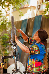 Using expert equipment, healthy young woman is repairing her own bicycle in yard as part of annual summer maintenance. Committed sporty african american lady fixes bicycle wheel with spanner as - obrazy, fototapety, plakaty