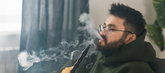 Banner bearded millennial or gen z man smoking hookah while relaxing on sofa at home copy space -...