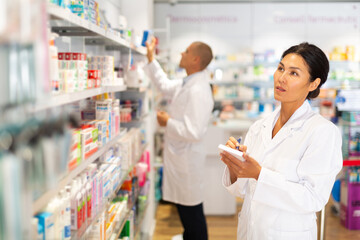 Asian woman in white gown checking medicines on shelves and comparing them with list. Her male...