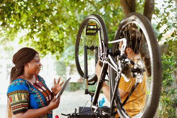 Vibrant black woman happily surfing internet to assist athletic man with bicycle maintenance....