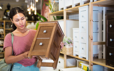 Asian woman holding small cabinet with two drawers in hands and looking in camera while standing in...