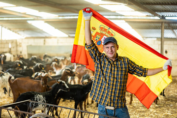 Happy young male traveler in plaid shirt waving flag of Spain during visit to livestock goat farm