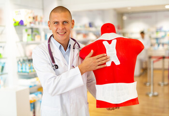 Male doctor standing in drugstore and holding red mannequin with medical bandages on it.
