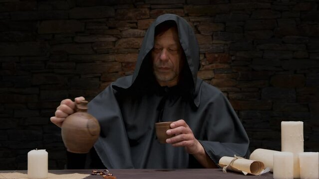 An old monk in a black robe pours wine into a goblet from a jug and drinks while sitting at a table against a stone wall