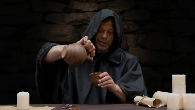 A monk in a black cassock pours and drinks wine while sitting at a table against a stone wall.