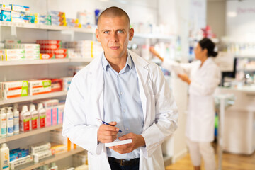 Doctor in white coat standing in salesroom with pan and recipe blanks in hands and looking in...