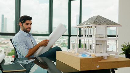 Smiling engineer looking project plan while design house model at office. Smart caucasian sitting...