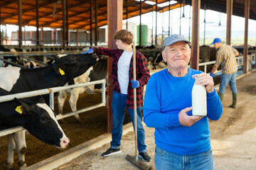 Portrait of smiling successful elderly farmer standing in cowshed at dairy farm, holding bottle of...