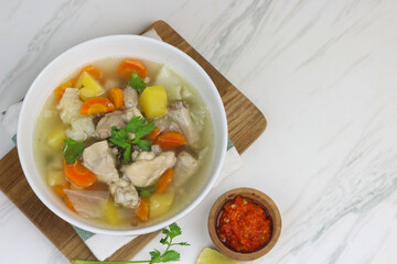 Chicken vegetables soup, Indonesian popular chicken soup, made from chicken meat, carrot, potato, and cabbage. served with sambal an lime slice. White marble background and copy space.