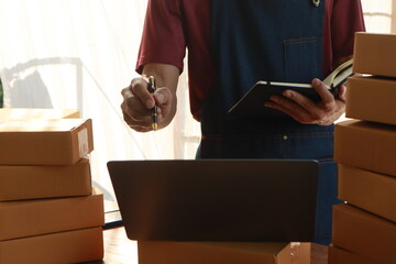Middle aged Asian man, the owner of a small business, happily works on her laptop at home. She's managing orders and packaging products for delivery, showcasing success in e-commerce.