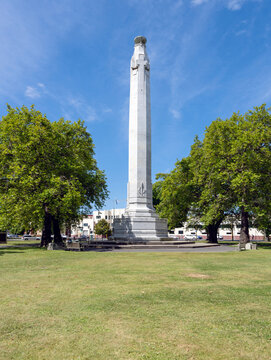 Dunedin, New Zealand - 9 September, 2023: Tall white obelisk war memorial featuring detailed sculptures at its base, set in a lush park with vibrant green trees and a clear blue sky.