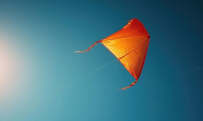 A red kite is flying in the blue sky. The kite is made of a thin orange fabric and has a long tail. The sun is shining brightly in the background.