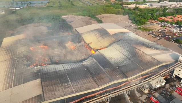 Intense firefighting rages at an industrial complex. Roaring flames breach the roof as multiple trucks douse them with torrents of water. A relentless battle against inferno unfolds.
