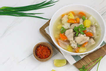 Chicken vegetables soup, Indonesian popular chicken soup, made from chicken meat, carrot, potato, and cabbage. served with sambal an lime slice. White marble background and copy space.