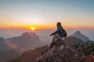 Adventurous Man Hiker with backpack sitting on top of steep rocky cliff mounting and enjoying sunset. Thailand