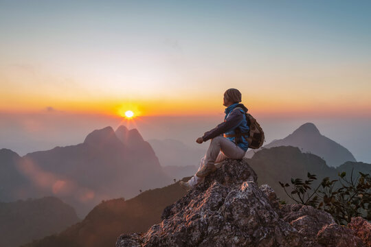 Adventurous Man Hiker with backpack sitting on top of steep rocky cliff mounting and enjoying sunset. Thailand
