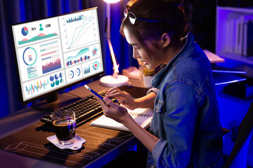 Thinking young beautiful Asian creative woman focusing on smartphone of sales target report business market graph comparing with report on computer at neon light modern office at night. Stratagem.