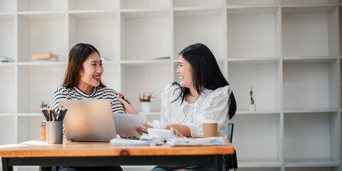 Two female colleagues enjoying a friendly chat with documents and a laptop on the desk in a spacious office setting.