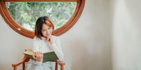 Introspective young woman engrossed in reading a book, seated comfortably by a circular window with...