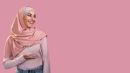 Woman recommending. Advertising background. Delighted cheerful girl in muslim headscarf presenting something invisible isolated on pink empty space.