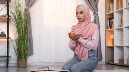 Koran praying. Faithful muslim. Islam culture. Calm focused woman in hijab with closed eyes reading Holy Quran at Ramadan on floor at light modern home interior with free space.