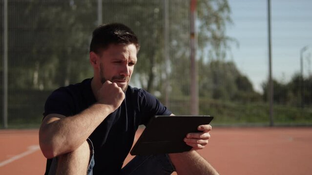 Caucasian man on sports field looks intensely at tablet and becomes emotionally upset. Sports fan is angry about team and his bad bet on bookmaker app. Portrait of bettor with negative emotions.