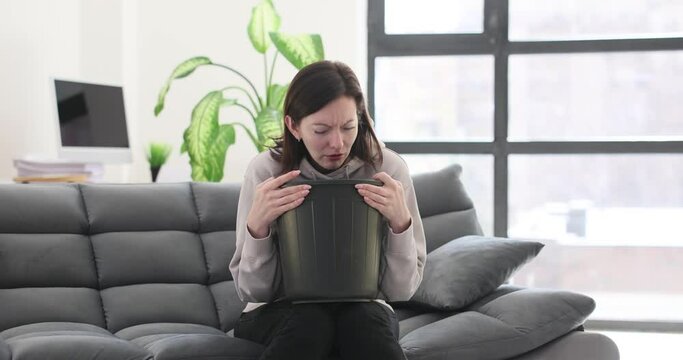 Young woman sits on a sofa with head in a trash can