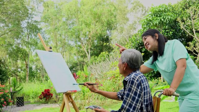 Asian nurse stand beside to take care near senior man sit on wheelchair and draw or paint for relax and revive good health of old people in outdoor with warm light.