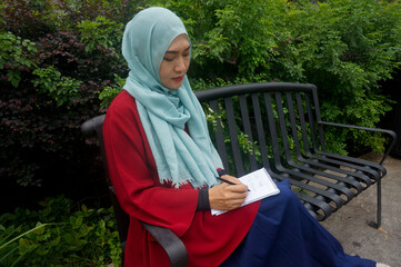 Asian muslim woman writing on notebook while sitting on the bench at the park