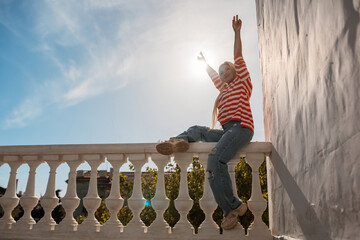 A woman is sitting on a railing with her arms raised in the air. The sun is shining brightly,...