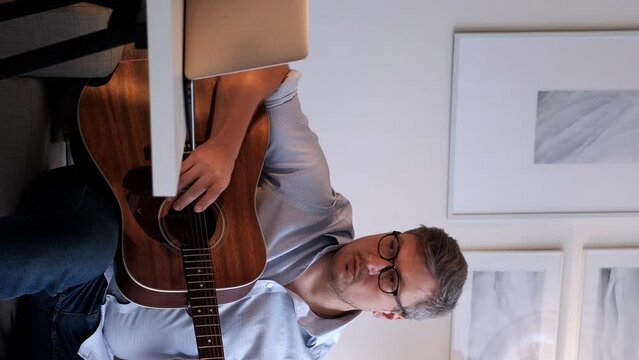 Vertical video. Music lesson. Chords practicing. Online meeting. Inspired professional man explaining rules playing acoustic guitar on laptop internet course sitting on sofa home.