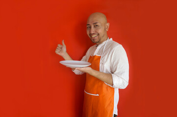 Young bald man wearing apron showing thumb up while holding empty white plate for mockup. Baker,...