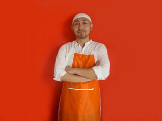 Portrait of adult Asian muslim man with skullcap and apron, standing in arms folded and looking at camera confidently