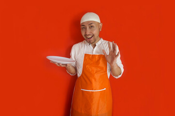 Adult Asian muslim man wear apron and cap, holding empty white plate mockup and showing happy...