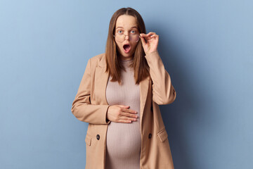 Shocked astonished young adult pregnant woman wearing dress and jacket posing isolated over blue...