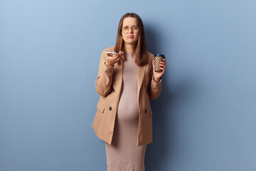 Unhappy dissatisfied young adult pregnant woman wearing dress and jacket holding mobile phone and...