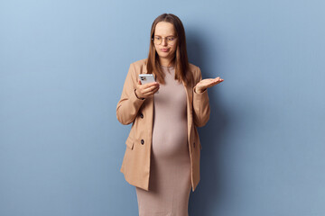 Confused puzzled young adult pregnant woman wearing dress and jacket holding mobile phone looking...