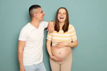Laughing positive cheerful pregnant Caucasian young couple standing isolated over light green...