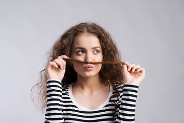 Portrait of a gorgeous teenage girl with curly hair, holding lock of hair as a moustache. Studio shot, white background with copy space