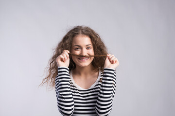 Portrait of a gorgeous teenage girl with curly hair, holding lock of hair as a moustache. Studio...