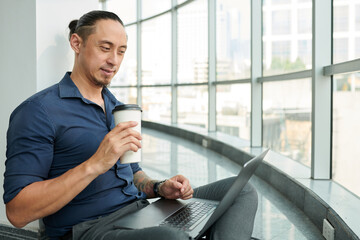 Smiling businessman drinking coffee and checking emails on laptop
