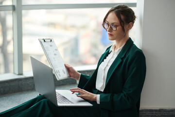 Serious businesswoman filling form on laptop with data from printed report