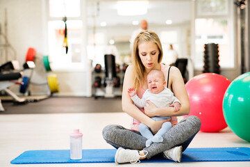 Portrait of new mom holding crying baby during group exercise class in gym. Moms staying active...
