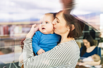 Portrait of new mom holding baby, standing by window in cafe. Spending free time during maternity...