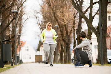 Overweight woman running in park, personal trainer checking her performance. Exercising outdoors...