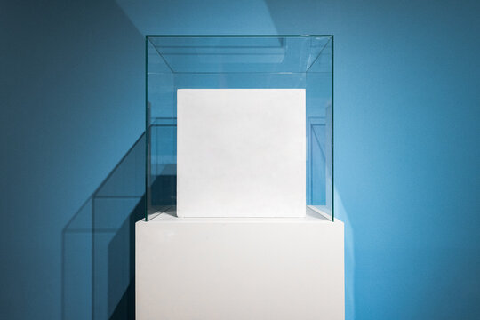Square white box mockups inside blank glass box podium mockup in exhibition gallery. Empty showcase mock up. Clear exhibition cube for museum or store. Display cabinet for expo.
