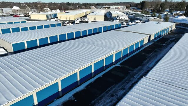 Self storage garage units covered in snow during winter. Aerial above rooftops with blue garage doors. Climate controlled rental space.