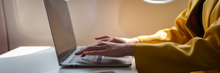 Fototapeta premium Young Asian business woman in yellow suit uses laptop sitting near window on airplane to do online financial business work Contact during flight, travel and business tourism concept.