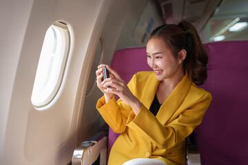 Young woman, business woman in a yellow suit is using a smartphone to take a photo of the scenery. Sky view from the window on an airplane with Wi-Fi internet connection. Travel business concept.