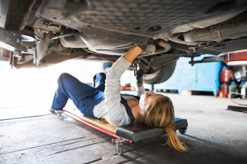 Female auto mechanic lying on mechanic creeper under car, inspecting and repairing vehicle. Beautiful woman working in a garage, wearing blue coveralls. - Powered by Adobe